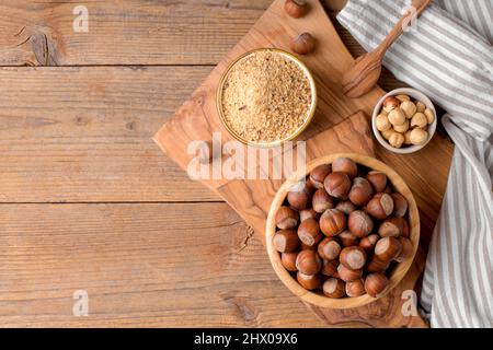 Whole raw hazelnut in a wooden bowl with peeled hazelnuts and nut flour on textured wooden background, top view Stock Photo