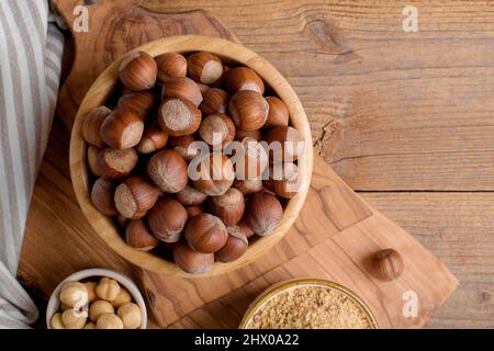 Whole raw hazelnut in a wooden bowl with peeled hazelnuts and nut flour on textured wooden background, top view Stock Photo