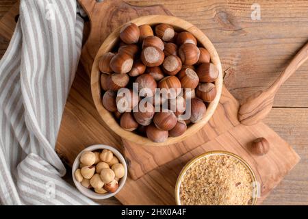 Whole raw hazelnut in a wooden bowl with peeled hazelnuts and nut flour on textured wooden background, top view Stock Photo