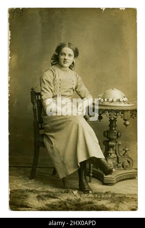 Original WW1 era postcard of pretty teenage girl, wearing a skirt with decorated blouse, and bow in her long hair, with decorated straw hat on the table. From the studio of Barcroft & Crabtree, with studios in Burnley and Nelson, Lancashire, England, U.K. circa 1914. Stock Photo