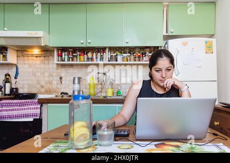 front view of young latin caucasian woman studying from home with her computer, attending online classes, staring at the computer with one hand on her Stock Photo