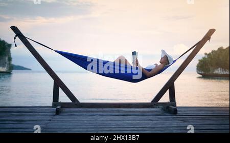 Shh, Im on vacation mode. Full length shot of an attractive young woman sitting alone in a hammock and reading a book during a vacation. Stock Photo