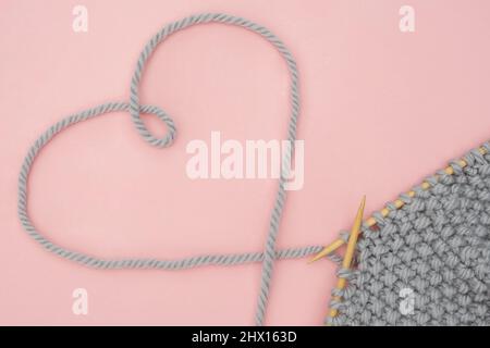 Piece of grey knitted fabric on wood needles and thread heart shape, process of knitting on pink background. Concept love for knitting, needlework. Co Stock Photo
