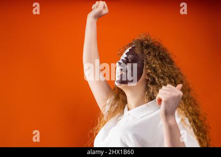Redhead fan football young woman celebrating her favorite football team success raising fists up with painted face in Qatar flag. Stock Photo
