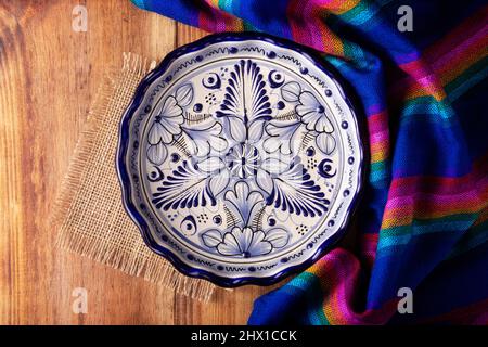 Mexican Cuisine Background, colorful traditional fabric and empty talavera style plate on wooden rustic table. Flat lay Stock Photo