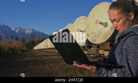 Woman student operator of institute of Solar Terrestrial Physics monitors communication equipment in notebook. Unique array solar radio telescope. Sun Solar Radio Telescope. The 'Quasar' observatory in Badary, Russia. Satellite dishes in mountains.