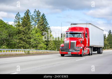 Clean well maintained big rig red semi truck tractor with truck driver rest compartment transporting cargo running on the straight wide highway road w Stock Photo