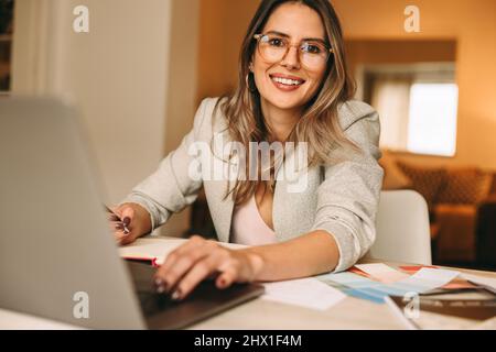 Happy female designer working in her home office. Creative businesswoman smiling at the camera while using a laptop at her desk. Cheerful interior des Stock Photo