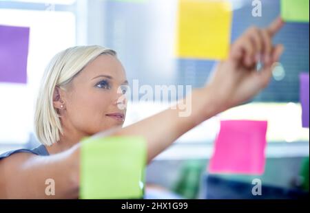 Planning her next move. Shot of a young woman in front of a transparent wall filled with post-its. Stock Photo