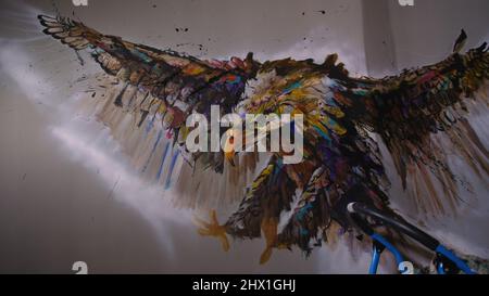 Artist designer draws an eagle on the wall. Craftsman decorator paints a picture with acrylic oil color. Painter painter dressed in a paint coat. Indoor. Dark magic cinematic look. Stock Photo