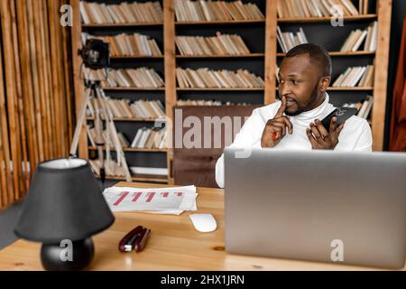 African american male freelancer or student recording voice message to a colleague, using smartphone. Successful businessman is working remotely from Stock Photo