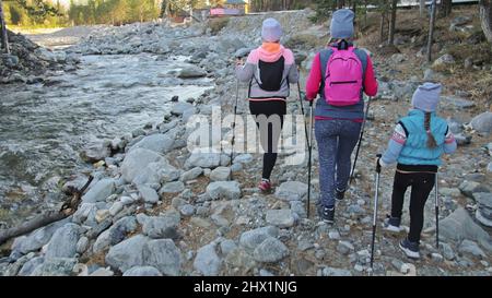 Woman do Nordic walking in nature near mountain river. Girls and children use trekking sticks and nordic poles, backpacks. Family travels sports. Kid is learning from mother and grandmother the proper technique of nordic walk. Autumn forest. Stock Photo