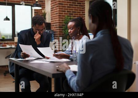 Marketing agency team members reviewing accounting documentation and revenue statistics data. African american business company colleagues in meeting about startup project financial status. Stock Photo