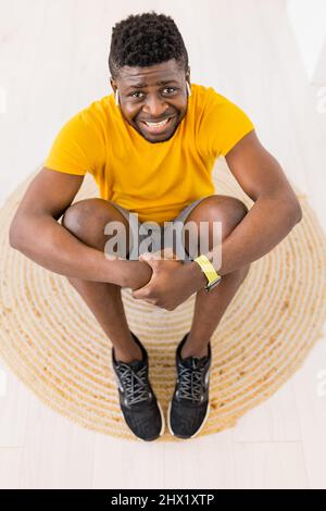 Handsome young African American male in yellow sportswear sitting on floor at home and embracing knee while smiling and looking at camera and resting Stock Photo