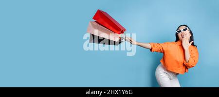 Young fresh and cheerful Asian woman in orange shirt holding and get shopping bag with big smiled on blue background, shopping on holiday and flash se Stock Photo