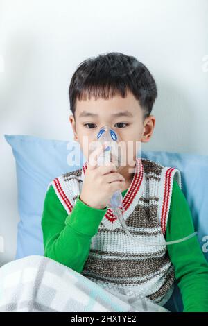 Sad asian child holds a mask vapor inhaler for treatment of asthma on sickbed in hospital. Breathing through a steam nebulizer. Concept of inhalation Stock Photo