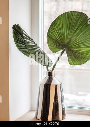Black and white stylish ceramic vase with green palm leaves on white windowsill. Front view. Copy space Stock Photo