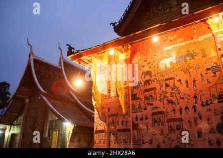 The exterior of the ancient Buddhist temple of Wat Xieng Thong at twilight. Spotlight shines on the stained glass on the wall. Luang Prabang, Laos. Stock Photo
