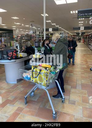 Lady paying for shopping in green coat and blonde hair with shopping trolley Waitrose Rushden Lakes Northamptonshire UK checkout paying mask Stock Photo