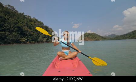 Attractive sportive blonde woman with sunglasses rows pink plastic canoe along sea water against green hills and blue sky. Traveling to tropical countries. Sports girl is sailing on kayak in ocean. Stock Photo