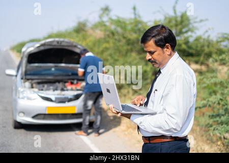 Businessman on roadside busy working on laptop in front of broken car while mechanic buy reparing car - concept of mobile car service, busy work Stock Photo