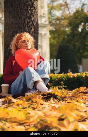 thoughtful young woman sitting under the tree autumn leaves holding red heart baloon - overall shot. High quality photo Stock Photo