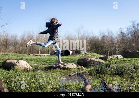 A young girl jumps dances over stones on an obstacle course in a park, in bright Spring sunshine, in a forest in Diemen, The Netherlands. Stock Photo