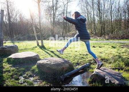 A young girl jumps between stepping stones on an obstacle course in a park, in bright Spring sunshine, in a forest in Diemen, The Netherlands. Stock Photo