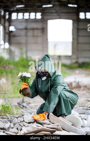 A man in a raincoat and gas mask collects a flower from a scorched, toxic land. Air pollution concept. Ecological catastrophy. Stock Photo