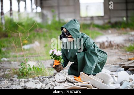 A man in a raincoat and gas mask collects a flower from a scorched, toxic land. Air pollution concept. Ecological catastrophy. Stock Photo
