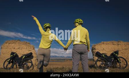 The man and woman travel on mixed terrain cycle bike touring with bikepacking. The two people journey with bicycle bags. Sport sportswear in green black colors. Mountain snow capped, stone arch. Stock Photo