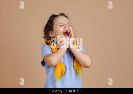 Shouting sad young girl with opened mouth touching face with hands crying having kanekalon braids of yellow colour on head wearing light blue t-shirt Stock Photo