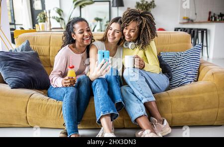 Three young multiracial women taking selfie while relaxing together at home Stock Photo