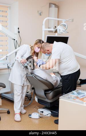 The dentist doctor looks at the patient's teeth and holds dental instruments near the mouth. The assistant helps the doctor. They wear white uniforms Stock Photo
