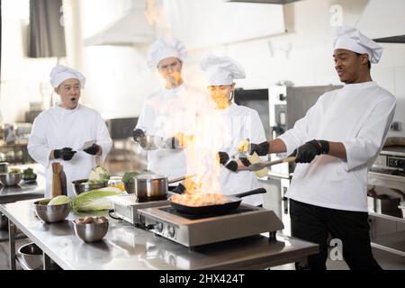 Multiracial team of professional cooks in uniform preparing meals for a restaurant in the kitchen. Latin guy burning pan, european cooks making sauce and asian chef managing the process. Teamwork and hard job at restaurant Stock Photo