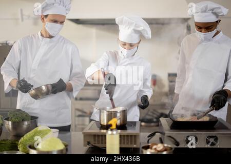 Multiracial team of cooks in uniform and face masks cooking meals for a restaurant in the kitchen. Concept of teamwork at restaurant during pandemic. Latin, Asian and European guys cooking together Stock Photo