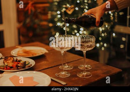 New Years Eve celebration. Man pouring champagne into glasses standing on table with festive xmas dinner, candles and wrapped gifts against blurred ba Stock Photo