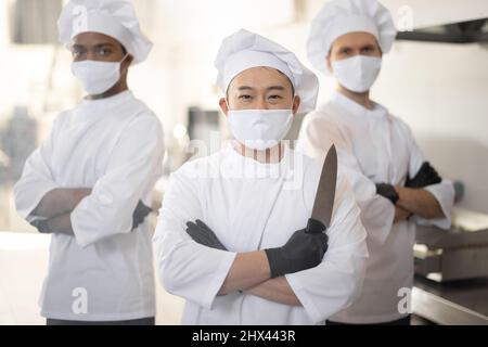 Portrait of three well-dressed chef cooks with different ethnicities standing together in restaurant kitchen. Asian chef holding knife, latin and european guys on background. Cooks wearing face masks ready to cook during pandemic Stock Photo