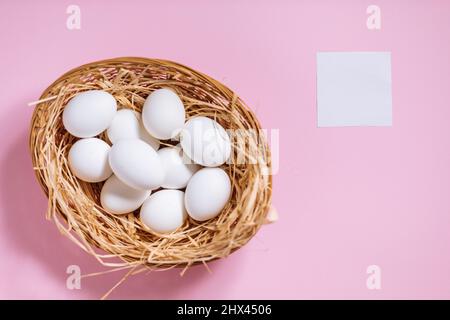 White easter eggs in yellow basket with straw on pink background. Top view of Happy Easter postcard concept Stock Photo