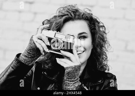 monochrome portrait of young beautiful cheerful woman with red curly hair taking pictures on film old fashioned camera on white bricks wall background Stock Photo