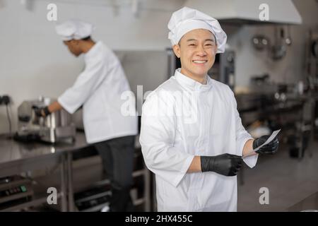 Portrait of smiling asian chef in uniform standing with printed order in professional kitchen with latin guy cooking on background Stock Photo