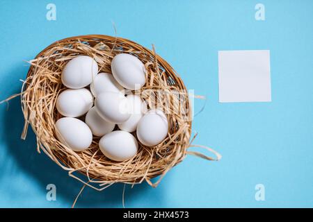 White easter eggs in yellow basket with straw on blue background. Top view of Happy Easter postcard concept Stock Photo