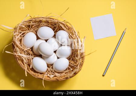White easter eggs in yellow basket with straw on yellow background. Top view of Happy Easter postcard concept Stock Photo