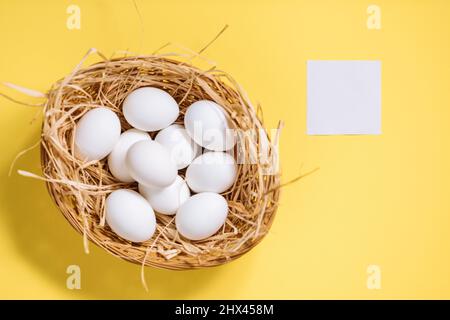 White easter eggs in yellow basket with straw on yellow background. Top view of Happy Easter postcard concept Stock Photo
