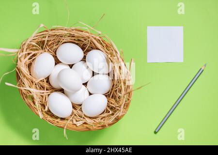 White easter eggs in yellow basket with straw on green background. Top view of Happy Easter postcard concept Stock Photo