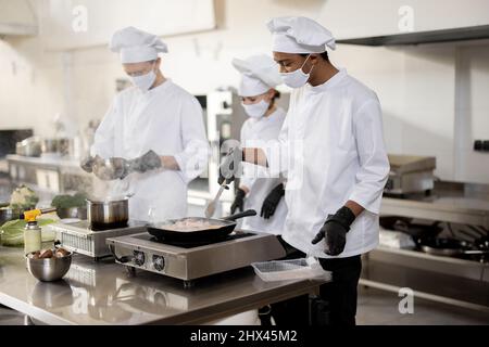 Multiracial team of cooks in uniform and face masks cooking meals for a restaurant in the kitchen. Concept of teamwork at restaurant during pandemic. Latin, Asian and European guys cooking together Stock Photo