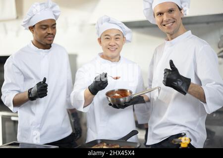 Portrait of three chef cooks with different ethnicities holding pan with a sauce while cooking in the kitchen. Teamwork and cooking delicious food at restaurant. Group of happy diverse cooks together at restaurant Stock Photo