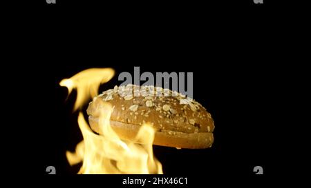 An appetizing burger bun with sesame seeds isolated on black background with red flame. Food and cooking concepr, rotating baked bun in flame. Stock Photo