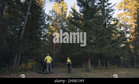 The man and woman travel on mixed terrain cycle touring with bikepacking. The couple journey with bicycle bags. Stylish bikepacking, bike, sportswear in green black colors. Magic forest park. Stock Photo