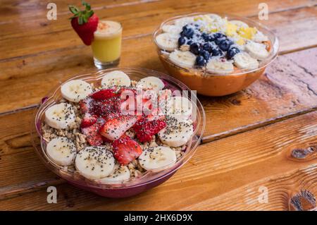 Two acai bowls with bananas and strawberries and a glass of juice with a fruit garnish on the natural pinewood table. Stock Photo